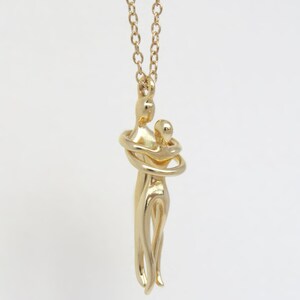 May include: Gold necklace featuring a pendant of two figures in an embrace. The figures are stylised and abstract, with the woman's arms wrapped around the man's neck.