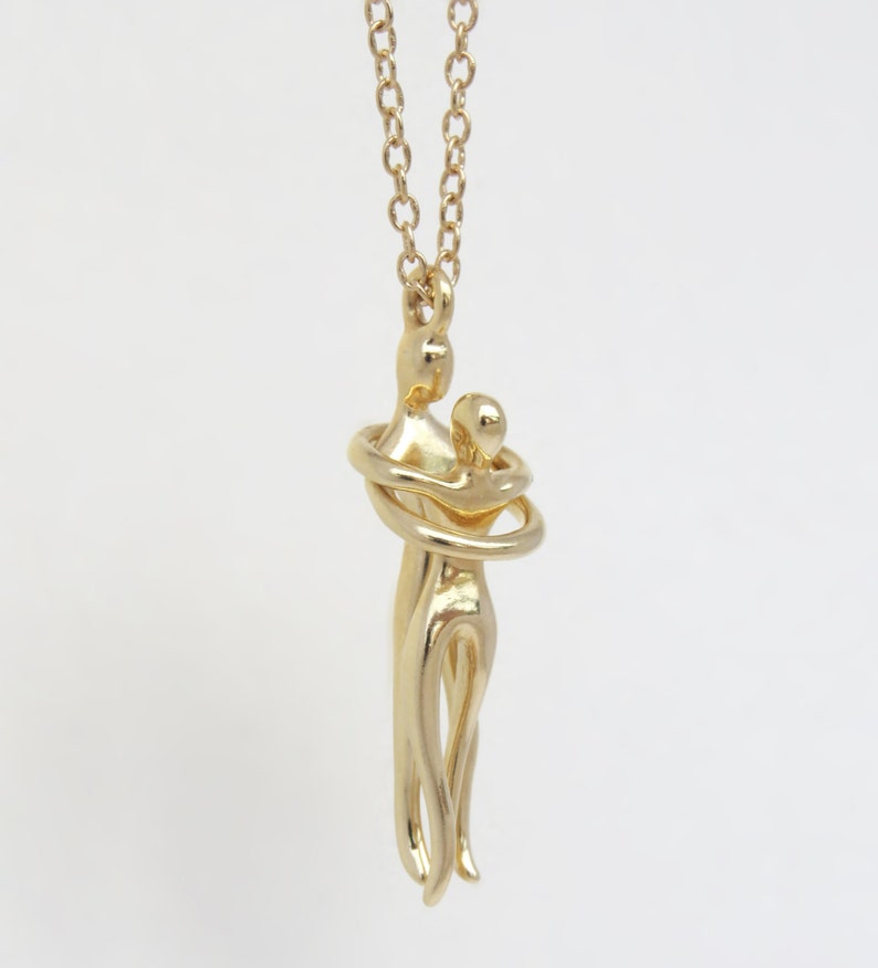 May include: Gold necklace featuring a pendant of two figures in an embrace. The figures are stylised and abstract, with the woman's arms wrapped around the man's neck.