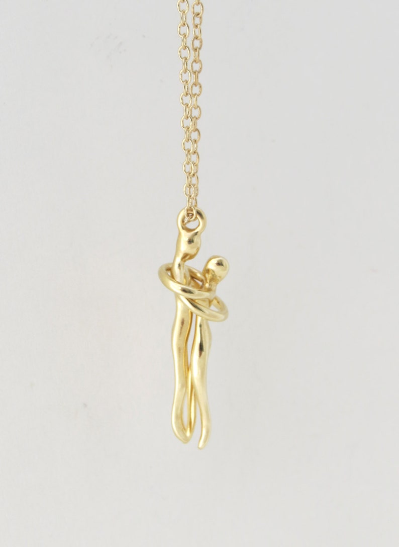 May include: A gold necklace featuring a pendant of two figures embracing. The figures are stylised and abstract, with the woman's arms wrapped around the man's neck.
