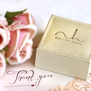 May include: A white jewellery box with a gold logo that reads "Sivan Lotan Jewellery". The box is sitting on a white surface with a pink rose bouquet and a thank you card.