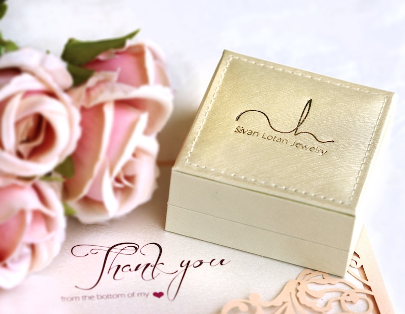 May include: A white jewellery box with a gold logo that reads "Sivan Lotan Jewellery". The box is sitting on a white surface with a pink rose bouquet and a thank you card.