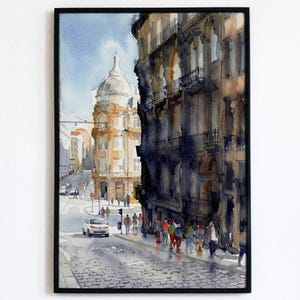 May include: Watercolor painting of a European city street scene. The artwork features a domed building and a tall building with balconies. People are walking on the cobblestone street. The painting is framed in a black frame.