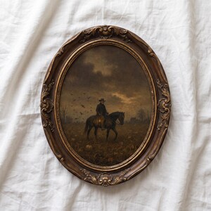 May include: An oval-shaped wall art piece featuring a dark, moody painting of a figure on horseback in a field under a cloudy sky, enclosed in an ornate, antique-style bronze-colored frame with intricate scrollwork and decorative floral details.