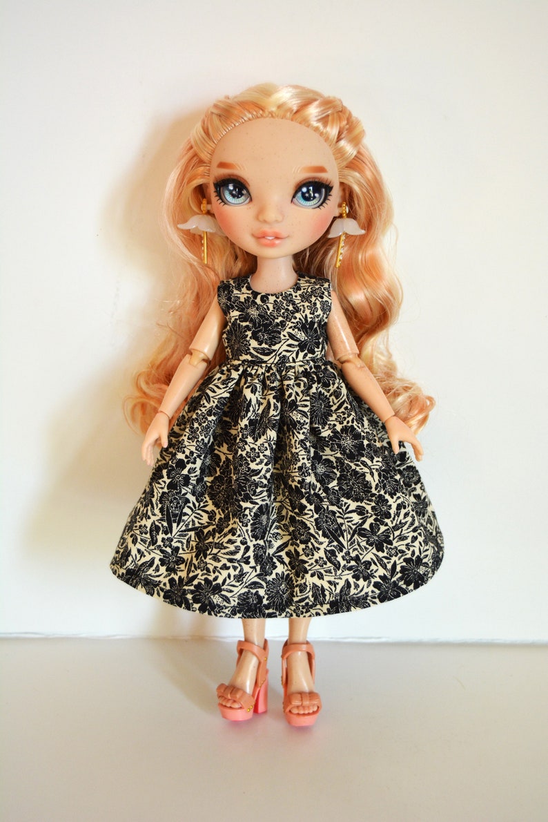 K&ouml;nnte beinhalten: Eine Puppe mit blonden Haaren, die ein schwarzes und cremefarbenes Blumenkleid und rosa High Heels tr&auml;gt.