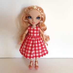 K&ouml;nnte beinhalten: Eine Puppe mit blonden Haaren, die ein rot-wei&szlig; kariertes Kleid und rosa Schuhe tr&auml;gt. Die Puppe hat blaue Augen und tr&auml;gt kleine wei&szlig;e Engelsfl&uuml;gel-Ohrringe.