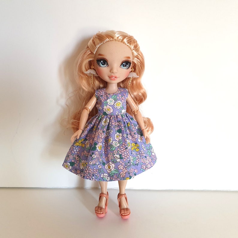 K&ouml;nnte beinhalten: Eine Puppe mit blonden Haaren, die ein lilafarbenes Kleid mit Blumenmuster und rosa Schuhe tr&auml;gt.