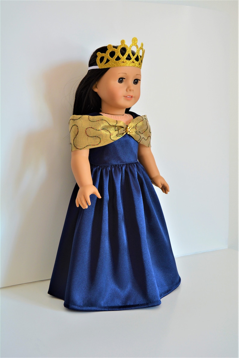 May include: A doll wearing a navy blue satin dress with a gold and red trim. The dress has a gold bow at the neckline and the doll is wearing a gold glitter crown.