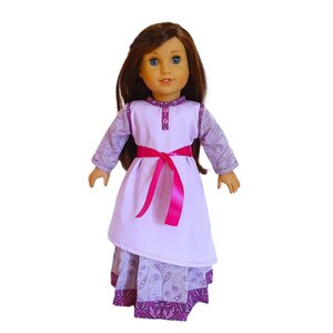May include: A purple and pink doll dress with a paisley print skirt and a solid purple bodice. The dress has a pink sash and a button closure at the neckline.
