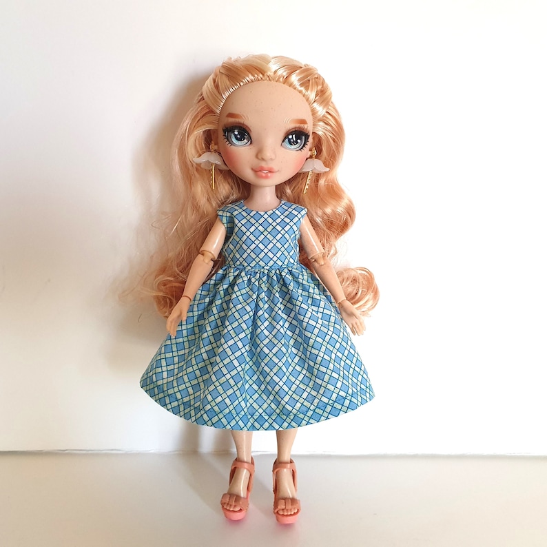 K&ouml;nnte beinhalten: Eine Puppe mit langen blonden Haaren, die ein blaues und gr&uuml;nes kariertes Kleid und rosa Schuhe tr&auml;gt. Die Puppe hat blaue Augen und tr&auml;gt wei&szlig;e Engelsfl&uuml;gel-Ohrringe.