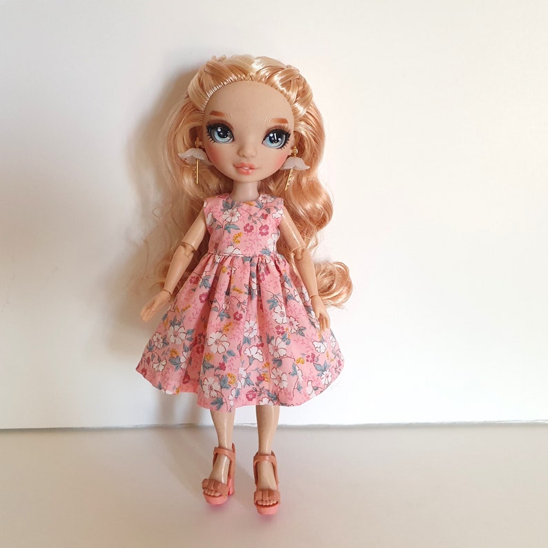 K&ouml;nnte beinhalten: Eine Puppe mit blonden Haaren, die ein rosa Blumenkleid und rosa Schuhe tr&auml;gt. Die Puppe hat blaue Augen und tr&auml;gt goldene Ohrringe.