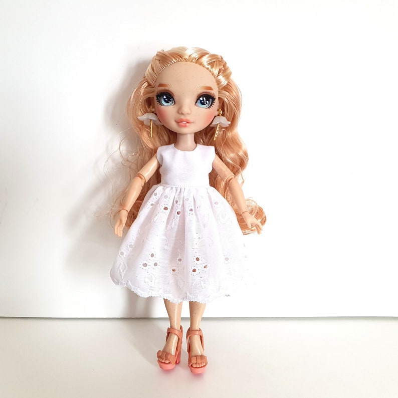 K&ouml;nnte beinhalten: Eine blonde Puppe, die ein wei&szlig;es Spitzenkleid und pfirsichfarbene Abs&auml;tze tr&auml;gt. Die Puppe hat blaue Augen und tr&auml;gt goldene Ohrringe mit wei&szlig;en Akzenten.
