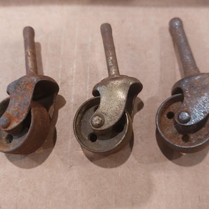 May include: Three antique metal casters with a rusty, aged appearance. Each caster has a small wheel, a curved yoke, and a long stem. The metal has a mix of brown and gold tones, suggesting age and wear.