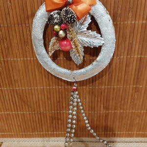 May include: Silver Christmas wreath with orange bow, pinecones, and red and gold ornaments. The wreath has a silver leaf and hanging silver beaded strands with two bell-shaped ornaments.