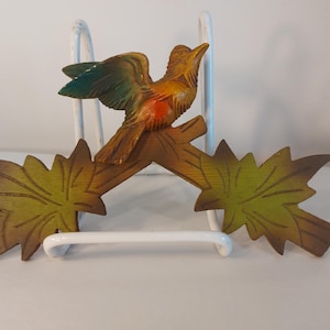 May include: A wooden bird figurine perched on a branch with green and yellow leaves. The bird has orange, brown, and green coloring. The figurine is set against a white background and supported by a white metal stand.
