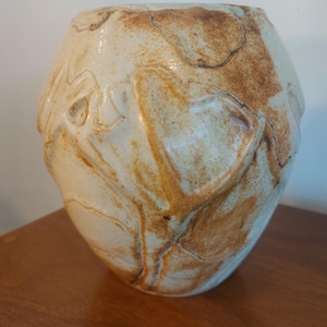 May include: A handcrafted ceramic vase with a unique, organic shape. The vase features a cream-colored base with brown, marbled accents and textured, leaf-like patterns. The vase is approximately 10 inches tall and 8 inches wide.