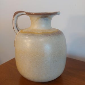 May include: A ceramic pitcher with a rounded body and a curved handle. The pitcher has a light beige base with brown and yellow accents around the rim and handle. The pitcher is likely used for serving beverages.