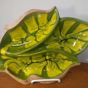 May include: A vintage ceramic serving dish with a leaf-like design. The dish features a vibrant color scheme of green and yellow, with a textured, glossy finish. The outer rim has a tan color. The dish is supported by a white metal stand.