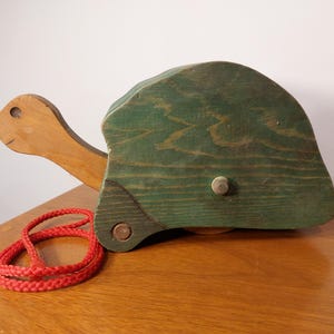 May include: A vintage wooden pull-along toy turtle with a green shell and a natural wood head and neck. A red braided pull cord rests beside the turtle. The toy is on a wooden surface.