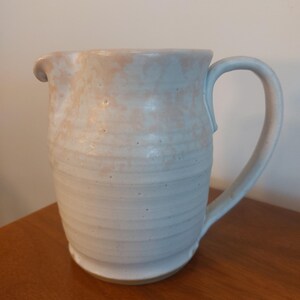 May include: A handmade ceramic jug with a light blue and peach glaze. The jug has a curved handle and a spout for easy pouring. The exterior features horizontal ridges, adding texture. This rustic jug is perfect for serving beverages.