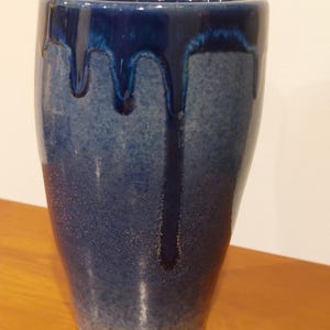 May include: A handcrafted ceramic vase with a gradient blue glaze. The top rim features a darker blue, with drips cascading down the sides. The vase has a cylindrical shape and a textured surface, perfect for home decor.