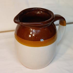 May include: A ceramic pitcher with a brown top and handle, a yellow stripe, and a white base. The pitcher has a spout for pouring and a curved handle. The overall design is simple and rustic.