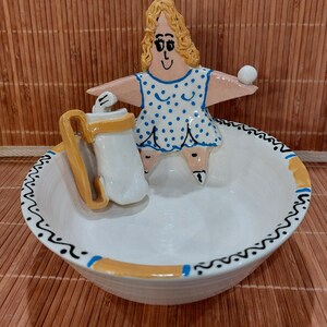 May include: A whimsical ceramic bowl with a hand-painted design. The bowl features a white base with a black and yellow patterned rim. A figurine of a woman in a blue polka-dot dress is in the center, holding a white ball. A white pitcher with a yellow handle is also present.