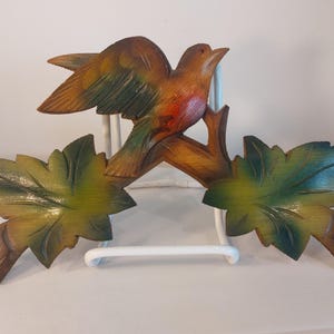 May include: A wooden bird sculpture perched on a branch with leaves. The bird has brown, orange, and green coloring. The leaves are green and yellow. The sculpture is mounted on a white metal stand.