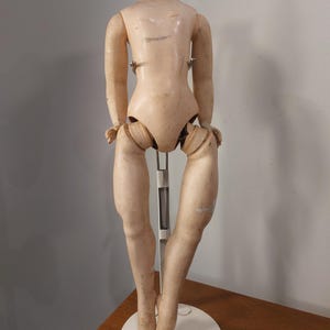 Peut inclure: Un corps de poupée vintage, de couleur chair pâle, est présenté sur un support en métal blanc. Les jambes de la poupée sont tendues et le corps est articulé. La poupée a un cou arrondi et une apparence lisse et vieillie.