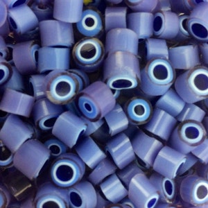 Op de afbeelding: Een close-up van talrijke lichtpaarse cilindrische kralen, elk met een donkerblauw en wit oogontwerp. De kralen zijn verspreid, waardoor hun cilindrische vorm en het gedetailleerde oogpatroon zichtbaar worden. Het oogontwerp heeft een donkerblauwe kern, witte en lichtblauwe ringen.