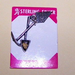 May include: Sterling silver charm of a pickaxe and shovel with a nugget of gold in the shovel. The charm is on a pink card with the text "Sterling Silver".