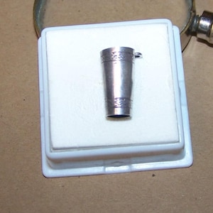 May include: A small, silver-colored pendant shaped like a miniature pint glass. The pendant has decorative etched bands near the top and bottom. It is displayed on a white surface inside a plastic case, with a magnifying glass in the background.