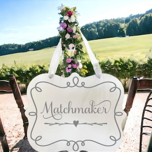 May include: White wooden sign with a gray border and the word "Matchmaker" written in cursive. The sign is hanging from a white ribbon and has a small gray heart and arrow design below the word.
