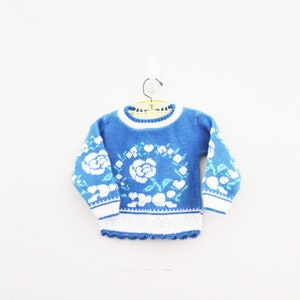 May include: A blue knitted sweater with a white floral pattern. The sweater has a white trim around the neckline and the bottom hem.