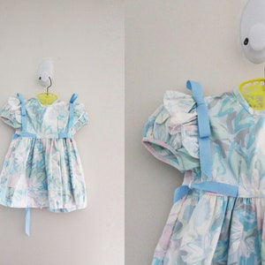 May include: A light blue and white floral dress with blue ribbon straps and a bow on each shoulder. The dress has a ruffled neckline and a gathered skirt.