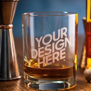 Könnte beinhalten: Ein klares Whiskyglas mit dem Text "YOUR DESIGN HERE" auf der Vorderseite, gefüllt mit bernsteinfarbener Flüssigkeit. Ein kupferfarbener Jigger und eine Flasche Bourbon befinden sich im Hintergrund, zusammen mit einem Korken.