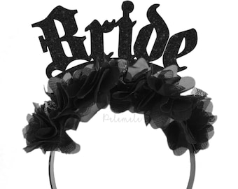 May include: A black headband featuring a row of black fabric flowers and a glittery black cutout sign that reads Bride in a stylized font. The accessory is designed to be worn on the head and displays a dark, monochromatic aesthetic throughout.
