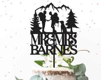May include: Black silhouette cake topper featuring a couple with backpacks hiking in the mountains with a dog. The text "MR & MRS BARNES" is below the couple.