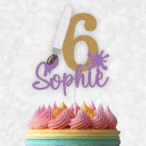 May include: Gold glitter number six cake topper with a purple glitter paintbrush and the name "Sophie" in purple glitter. The cake topper is on top of a rainbow layered cake.