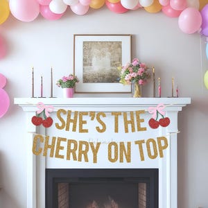 May include: A decorated fireplace with a gold glitter banner saying "SHE'S THE CHERRY ON TOP." The scene is adorned with colorful balloons, flowers, and decorative items. The setting suggests a party or celebration, with a framed picture and other details.
