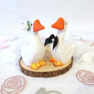 May include: Two white goose figurines with orange beaks and feet standing on a circular wood slice. One goose wears a white floral veil, and the other wears a black bow tie with a small pearl accent. The text www.pelemele.etsy.com is at the top.