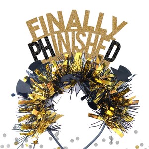 May include: Gold and black headband with the words "FINALLY FINISHED" in glitter letters. The headband is decorated with black and gold tinsel.