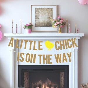 以下が含まれることがあります： 暖炉の上に、「A Little Chick Is On The Way」と書かれた金色のグリッターバナーが飾られています。バナーには、黄色いひよこのシルエットが描かれています。