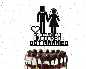 May include: Black and white cake topper with a pixelated couple and the text "Achievement Unlocked Got Married". The topper is on a chocolate cake with white frosting and chocolate drizzle. The cake is decorated with chocolate sprinkles and the name "Felemele" is written in gold glitter on the side.