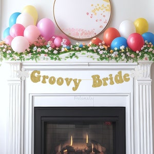 May include: A gold glitter banner with the text "Groovy Bride" hanging above a white fireplace mantel decorated with pastel balloons and greenery.