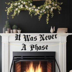 May include: Black and white banner with the text "It Was Never A Phase" hanging above a fireplace. The banner is made of paper and has a gothic font. The fireplace is white and has a fire burning inside.