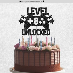 May include: Black glitter cake topper with a video game controller design and the text "LEVEL +8 UNLOCKED".