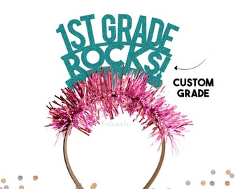 May include: Teal glitter headband with the text "1st Grade Rocks!" and a pink tinsel trim. The headband is customizable with a different grade.
