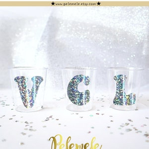May include: Three clear plastic shot glasses with silver glitter letters spelling "VCL".