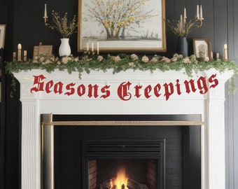 May include: White fireplace mantel with a garland of greenery and white flowers. The text "Seasons Creepings" is displayed in red on the mantel. The fireplace has a black surround and a lit fire inside.