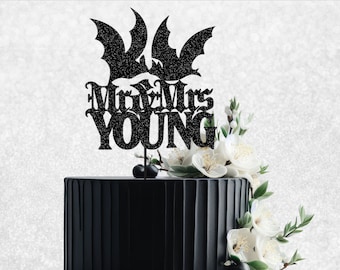 Può includere: Topper per torta nero con due pipistrelli e il testo "Mr & Mrs YOUNG" in un font gotico. Il topper è su una torta nera con fiori bianchi.